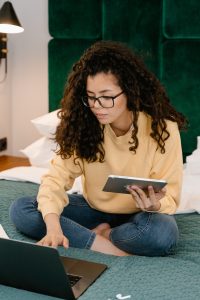 Woman works from home, sitting on bed holding a tablet while typing on a computer with the other hand.